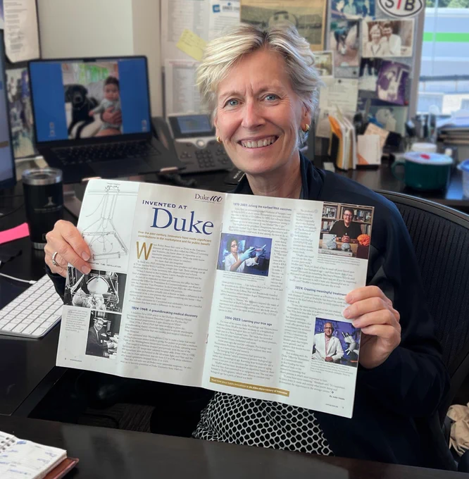 Robin Rasor sits smiling at her desk, holding up a Working@Duke magazine to the camera open to a two page spread entitled "Invented at Duke".