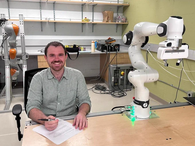 Preston sitting at a table with two robots, signing their offer letter from @Cornell_CS.