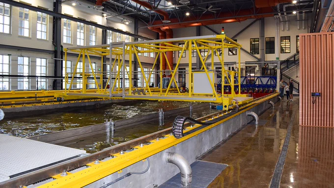 Interior of a water research facility featuring a large, yellow mechanical structure spanning over water basins, with surrounding industrial equipment and pipes.