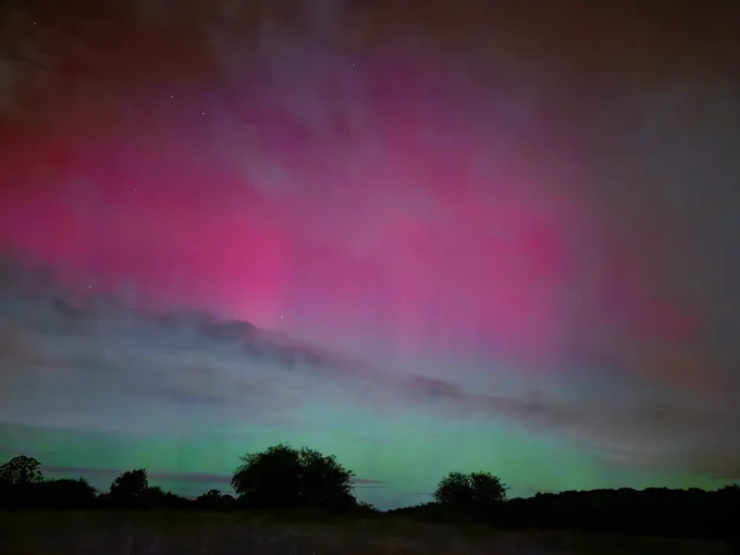 An image of the aurora across the sky in South Wales. The aurora is above a line of trees and hills.