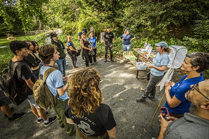 A group of individuals in casual attire gathers in a lush green park listening to someone standing beside a table with informational materials, maps, and snacks. A few attendees are standing, while one is seated in a folding chair.