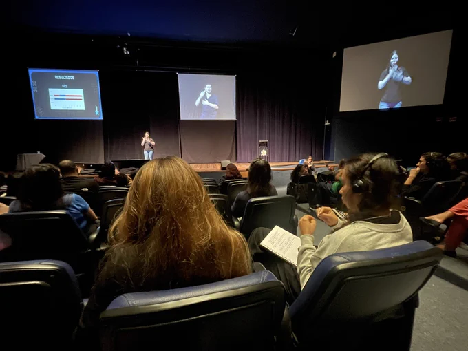 auditorium where a presentation is taking place. The speaker is on stage with a screen to the left displaying a slide titled “RESULTADOS” with a chart. Another screen to the right shows an interpreter. The audience is seated, with some wearing headphones.