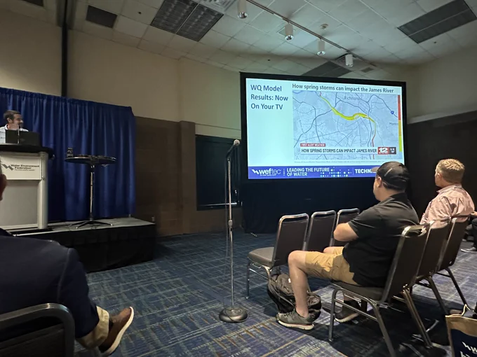 This picture showcases a speaker on a stage at a podium at the far left edge of the photo, and a presentation projected onto a screen on the right. The slide features a large screenshot of an NBC12 On Your Side story titled “How Spring Storms Can Impact the James River” that features the water quality model. The slide is titled on the left and is called “WQ Model Results: Now On Your TV.” “WQ” stands for “Water Quality.” As the photo was taken from the middle of the room in the third row, just three attendees are seen sitting in audience in the foreground of this photo in the second row on both sides of the aisle—one on the left and two on the right. There is a microphone on a stand at the front of the aisle and patterned, primarily blue carpeting in the room. A blue curtain hangs behind the stage and speaker, and a small black table sits on the stage between the podium and the screen. The Water Environment Federation logo decorates the front of the podium.