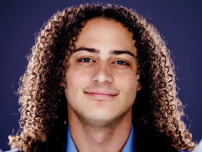 Portrait of a man with shoulder-length curly hair and a smile, wearing a light blue shirt against a dark blue background.