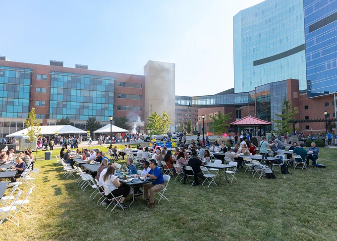 Outdoor event at a corporate campus with people seated at long tables, enjoying food and conversation. A modern glass building is visible in the background.