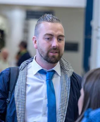 Josh Feehan with a beard and moustache, wearing a coat over a white shirt and blue tie, with a bag slung over his shoulder.