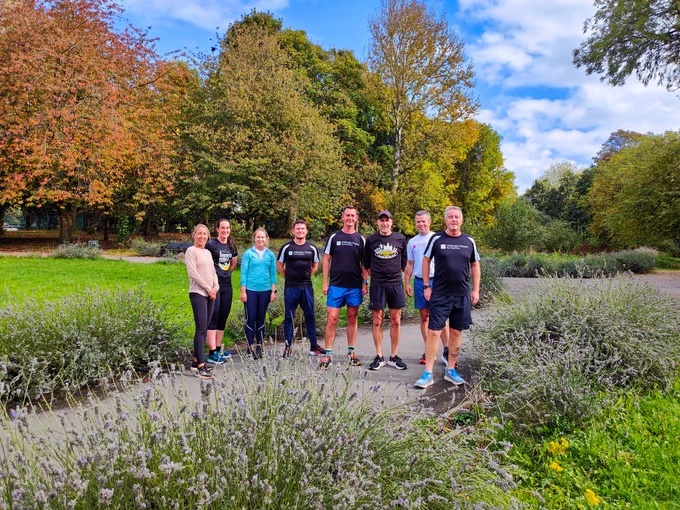 Seven runners stood in a line facing the camera in the greenery of Burley Park.  Lavender bushes in the foreground with autumnal fading purple flowers.  Trees turning orange and red in the background.  Blue skies with white clouds.