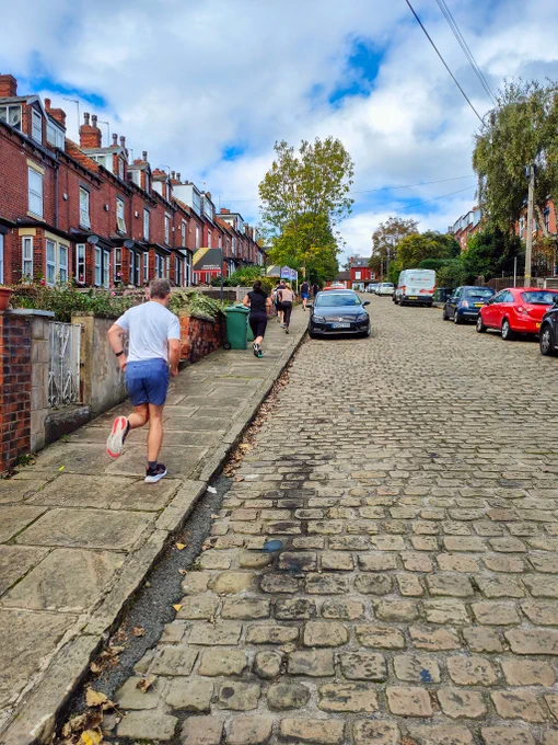 Runners running away from the camera up hill on a stone pavement.  The adjacent road is cobbled and red brick terrace houses line the street.  Blue skies with white clouds.