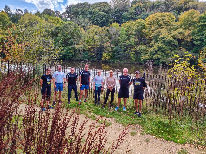 Seven runners stood in a line facing the camera next to the River Aire.  Trees turning orange but mainly green in the background.  A river wharf is just behind the runners, with a fence in between. Blue skies with white clouds.