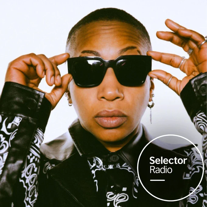 Press photo of OneDa, close up on her face wearing sunglasses and black leather jacket. On top of the image is the circular Selector Radio logo in the bottom right hand corner.