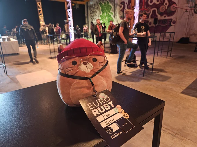 A beaver plushie with glasses and a EuroRust conference badge with name "Fish" and stickers "skilled user", "beaver", "10x engineer" and "job hunting".