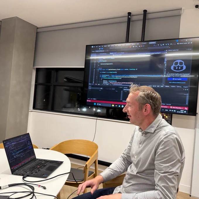 Lasse Fredslund sitting at a laptop which is projected on a monitor showing Visual Studio and the source code of a Win Forms application