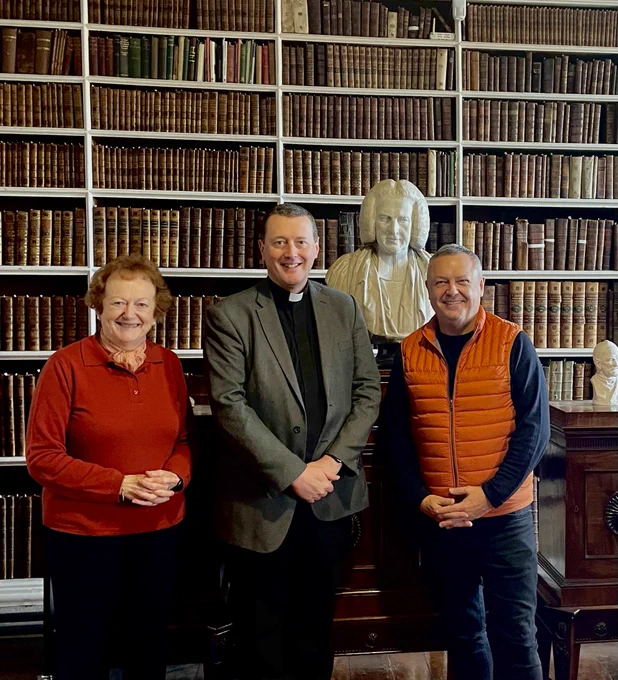 Three people, smiling, standing in front of bookshelves with old books, and a bust.