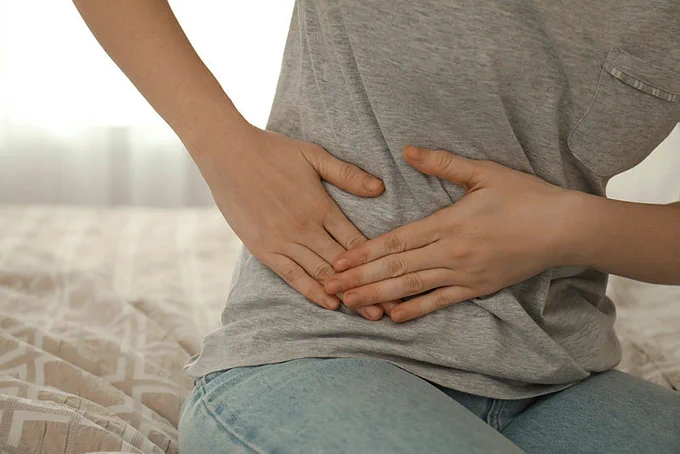 Woman holding their abdomen suffering from appendicitis pain sat on a bed at home, closeup. © New Africa / stock.adobe.com