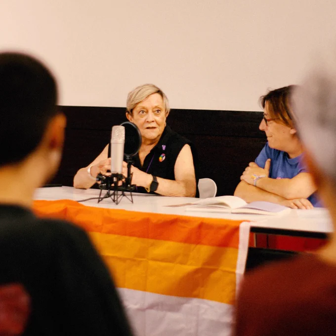 Guillermina, nunha foto do faladoiro durante o Orgullo PTV , organizado por Avante LGBT  Pontevedra. Deste evento saiu o podcast que podes escoitar na ligazón do tweet. Guillermina conversa coa Eva Mejuto nunha mesa decorada pola bandeira lésbica, e cun micro diante. Nun primeiro plano, borrosas, algunhas siluetas do público.