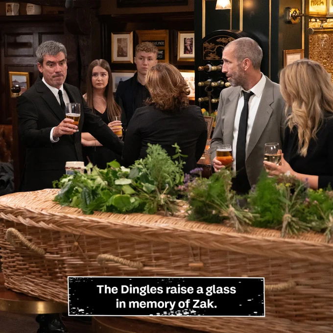 The Dingle raises a glass in the Woolpack. Title reads: The Dingles raise a glass in memory of Zak.
