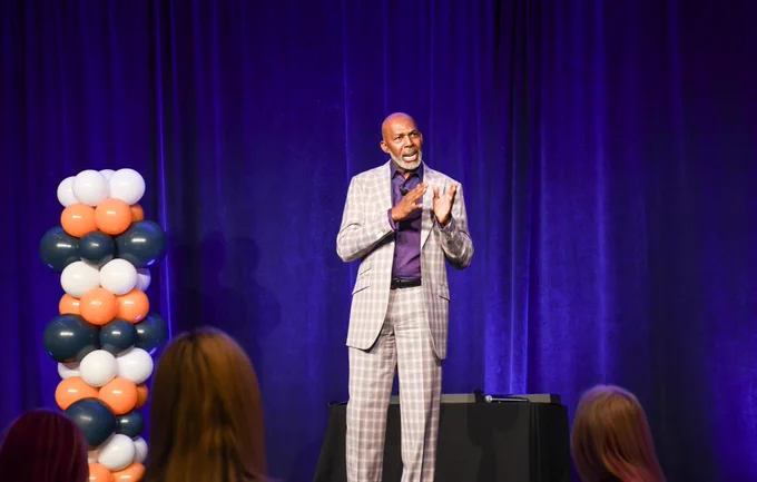 Thurl Bailey – Former Utah Jazz basketball player, NCAA champion, author & commentator – took the stage at The Grand America Hotel, speaking to members of the HCA Healthcare Mountain Division Leadership Institute (LDI) about Championship Leadership. Thurl, wearing a grey plaid suit with a purple dress shirt underneath, stands center and a vertical balloon arch is situated behind him with horizontal stripes of blue, orange and white balloons.