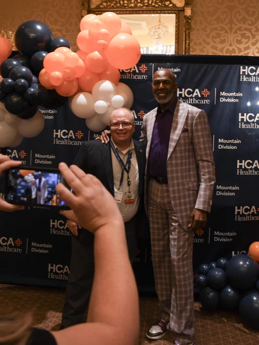 Thurl Bailey – who spoke at the HCA Healthcare Mountain Division Leadership Institute (LDI) in Salt Lake City, Utah – stands next to a colleague from Ogden Regional Medical Center, posed and smiling, in front of a branded step and repeat backdrop for a photo opportunity. Blue, orange and white balloons are also attached to the backdrop. The photo shows someone taking the exact photo on their cell phone, as well.
