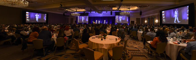 Thurl Bailey – Former Utah Jazz basketball player, NCAA champion, author & commentator – took the stage at The Grand America Hotel, speaking to members of the HCA Healthcare Mountain Division Leadership Institute (LDI) about Championship Leadership. Thurl, wearing a grey plaid suit with a purple dress shirt underneath, stands center and his video image is projected on numerous large screens in the ballroom.
