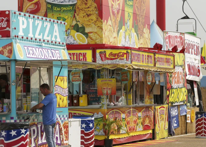 A colorful row of food stands at the Mississippi State Fair, with offerings such as pizza, lemonade, and corn dogs. Banners feature logos of snacks. A person is visible, attending one of the stands.