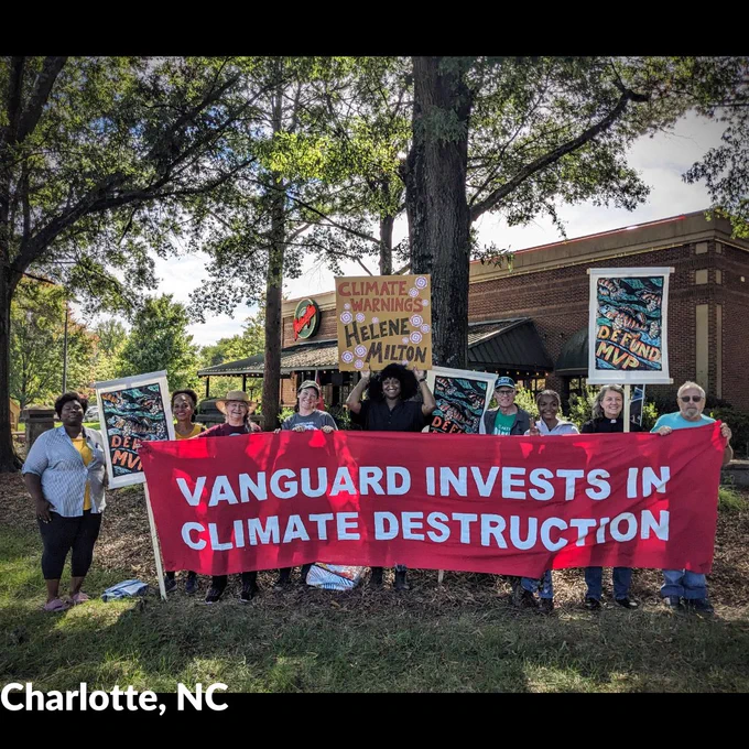Nine people stand holding various banners and signs, including one large red banner with white text. Photo by Josh Vana.