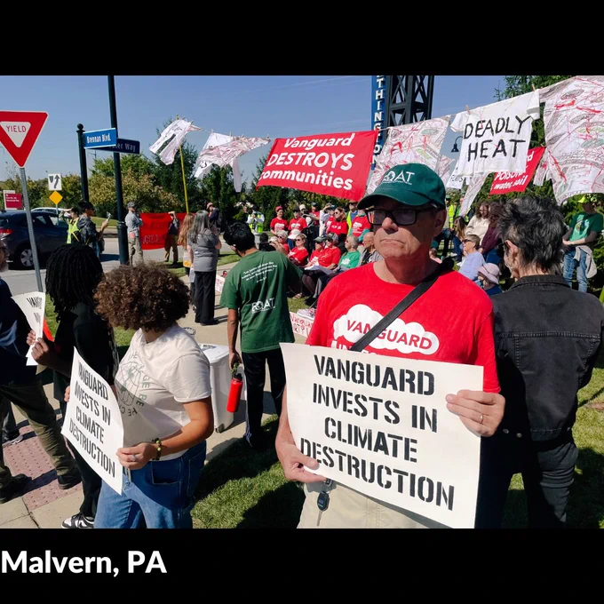 A few people stand, holding white signs with black text. Behind them are many more people sitting and standing and various banners and props. Photo by Rachael Warriner.