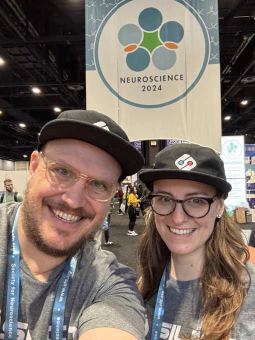 Two SilicoLabs team members smile for a selfie at the Society for Neuroscience 2024 conference, standing in front of the event banner while wearing matching branded hats and lanyards.