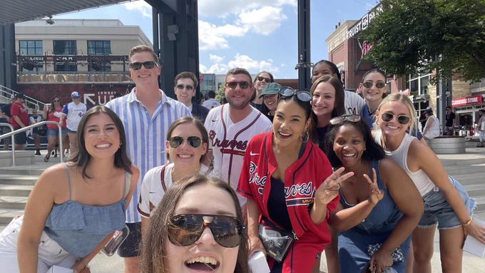 A group of smiling people at a sports event, some wearing Atlanta Braves jerseys, gathered for a selfie under a clear sky.
