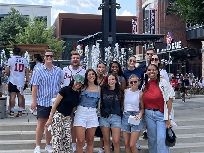 A group of people smiling in front of The Battery Atlanta near Truist Park's main gate. They are casually dressed, enjoying a sunny day outdoors.