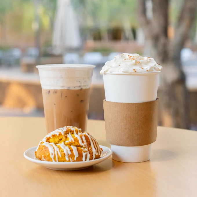 A pumpkin spice latte, an iced chai topped with pumpkin cold foam, and a pumpkin scone on a table. MIM’s Main Courtyard is in the background.