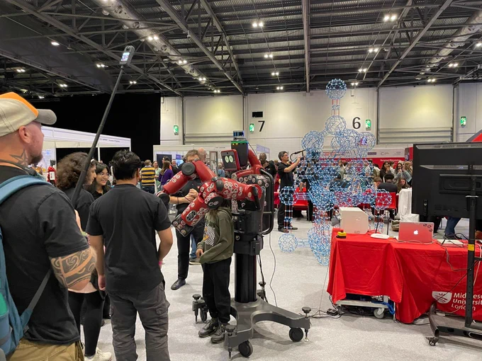Attendees at a tech expo examining a robotics display featuring a large red robotic arm, with various booths and exhibitors visible in the background.