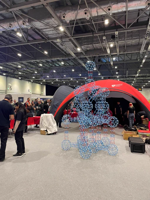 A vibrant promotional event featuring a large red dome. In the foreground, a decorative sculpture made of blue lights is displayed, surrounded by attendees in a busy exhibition hall.