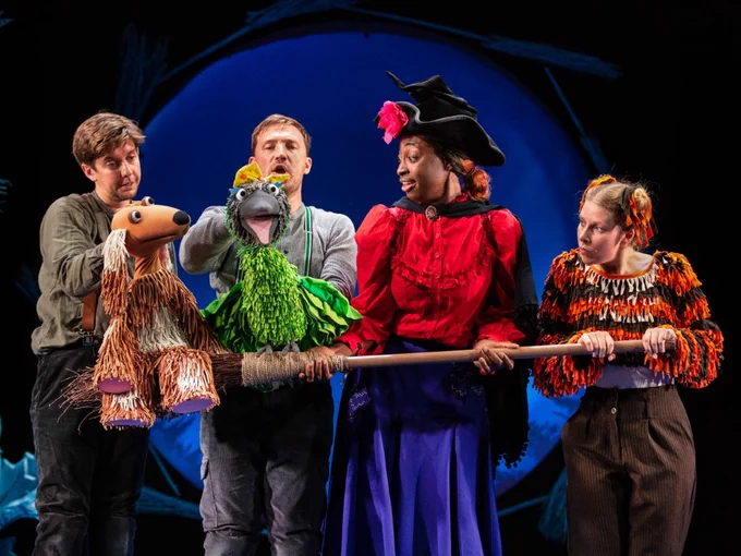 Four performers on stage holding puppets in a theatrical production. One performer wears a hat resembling a witch, set against a blue-lit background.