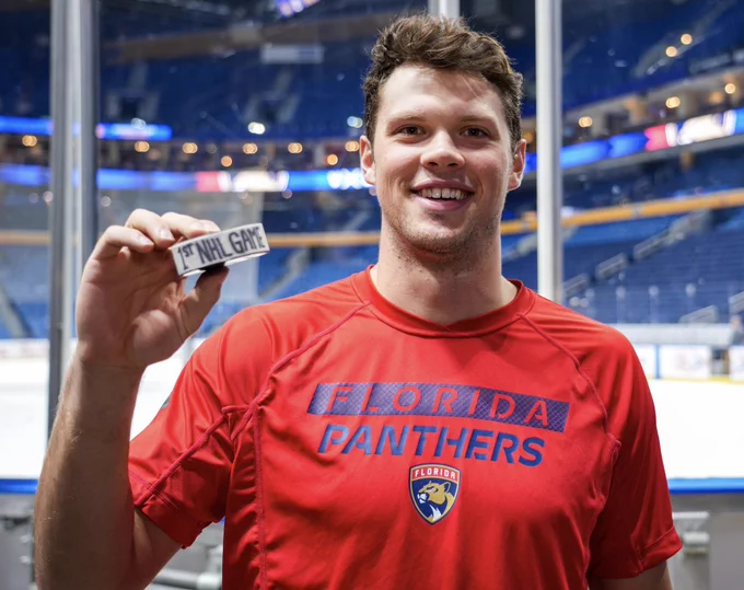 Panthers forward Patrick Giles smiles while posing with his first NHL game puck.

He is wearing a red Panthers t-shirt.
