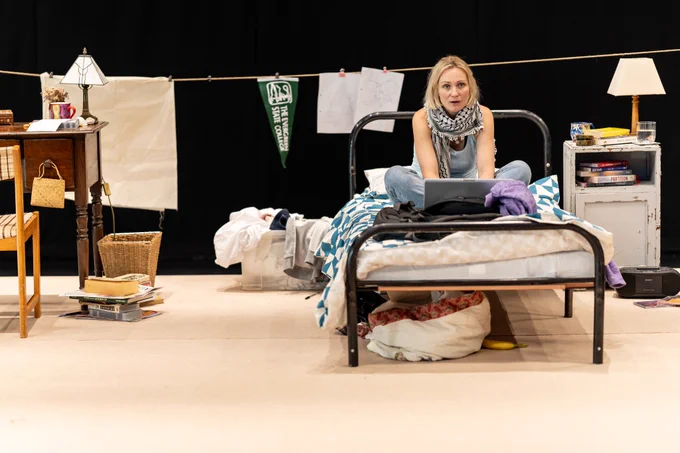 Rehearsal photo for Fy Enw i yw Rachel Corrie - a blonde white woman sits on an untidy bed with an open laptop in front of her. She's wearing a keffiyeh scarf.