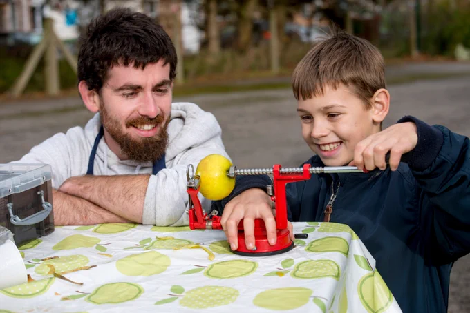A man watches as a boy uses an apple peeler. 