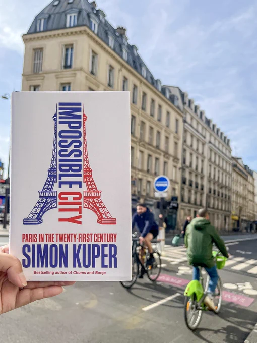 A hand holds a hardcover book: "Impossible City" by Simon Kuper, in front a building and bike lane in the center of Paris.
