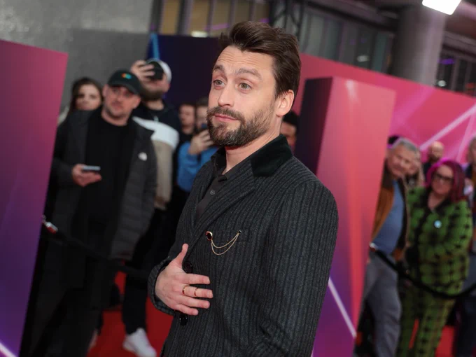 Kieran Culkin at a red carpet event wearing a dark striped suit and smiling at the camera.