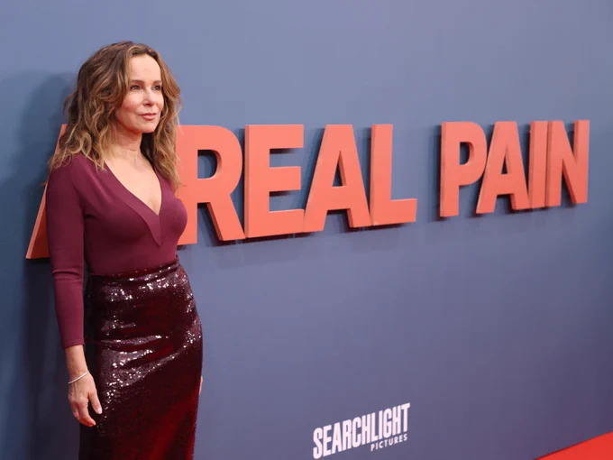 Jennifer Grey standing in front of a backdrop bearing the text A Real Pain, wearing a maroon top and a sequinned skirt.