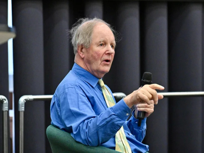 A man wearing a blue shirt sits on a stage and holds a microphone. He is answering questions from an audience.