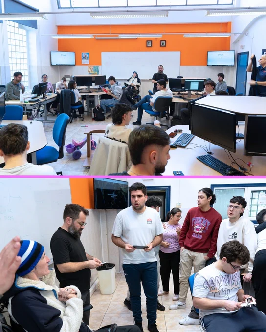 Grupo de personas en un aula, algunas sentadas y otras de pie, alrededor de mesas con ordenadores, mientras una persona en silla de ruedas eléctrica participa en la actividad.