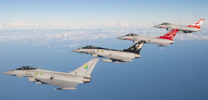 Image of four RAF Typhoons in assorted squadron colours flying in formation through cloudy light-blue skies.