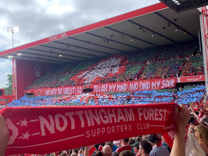 Photo of a packed Trent End stand at Nottingham Forest’s City Ground Stadium. The image is facing diagonally right from within the Brian Clough stand and at the foot of the picture, taking up the bottom third is a red scarf with Nottingham Forest written in white from left to right. There is a colourful display shown in the Trent End stand with the top of the Forest crest (a tree) depicted in the upper tier of the stand. The bottom tier is predominantly blue whilst a banner stretches the width of the stand directly between both tiers, the wording on this banner is as follows: “And into the Forest I go, to lose my mind and find my soul!”