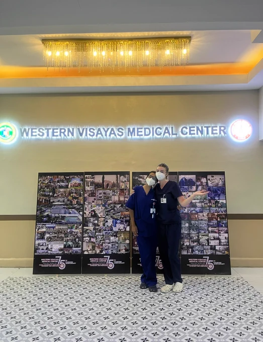 Two nurses in blue scrubs and PPE masks pose underneath a glowing sign that says Western Visayas Medical Center