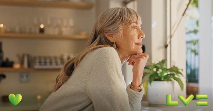 An older lady is in her kitchen, looking out of the window looking contemplative. 