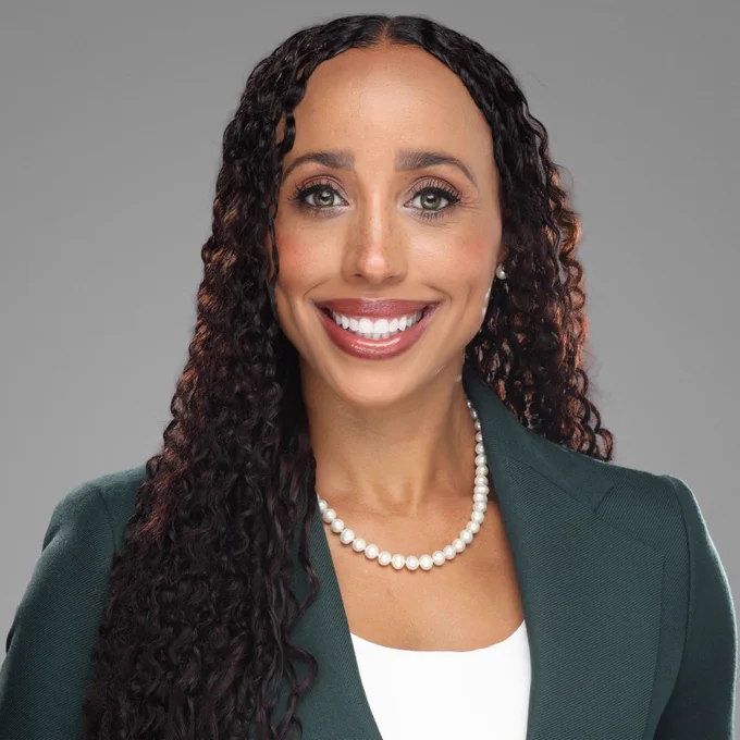 A woman with long curly dark hair smiling at the camera, wearing a dark green blazer over a white top and a pearl necklace, posed against a light gray studio background.