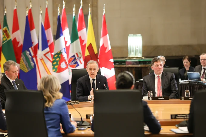 A group of officials sit around a conference table in front of multiple provincial and Canadian flags, engaged in a formal meeting. The focus is on a man speaking at the center, with others listening attentively.
--
Un groupe de fonctionnaires est assis autour d'une table de conférence, devant plusieurs drapeaux provinciaux et canadiens, et participe à une réunion officielle. L'accent est mis sur un homme qui parle au centre, les autres écoutant attentivement.