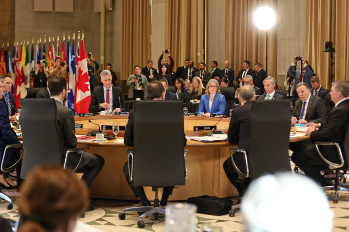 A group of officials sit around a large conference table in a formal meeting room, with national flags in the background and several photographers and media capturing the event.
--
 Un groupe de fonctionnaires est assis autour d'une grande table de conférence dans une salle de réunion formelle, avec des drapeaux nationaux en arrière-plan et plusieurs photographes et médias capturant l'événement.