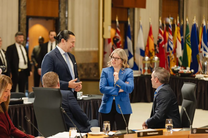 People in formal attire converse in a conference room with international flags in the background. A woman in a blue blazer stands talking to a man in a suit, while others sit and observe.
--
Des personnes en tenue formelle discutent dans une salle de conférence avec des drapeaux internationaux en arrière-plan. Une femme vêtue d'un blazer bleu discute avec un homme en costume, tandis que d'autres s'assoient et observent.