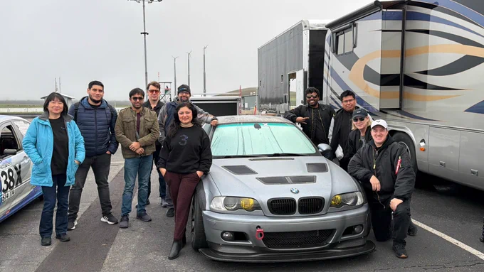 A group of ten smiling individuals pose around a silver BMW race car on a paved track under an overcast sky. The car features a BMW logo on its grille, custom hood vents, yellow headlights, a red tow hook, and a small "B" sticker on its front bumper; a person in the foreground wears a black sweatshirt with a prominent glittery number "3". In the background, a white race car with "38X" and "NASA" text is visible, alongside large support vehicles and tall wind turbines.